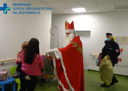 św. Mikołaj na Oddziale Dziecięcym wręczający pacjentowi prezent, w tle widoczny policjant niosący prezenty