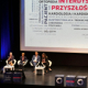 Cztery osoby siedzące w fotelach na scenie podczas konferencji. W tle ekran.