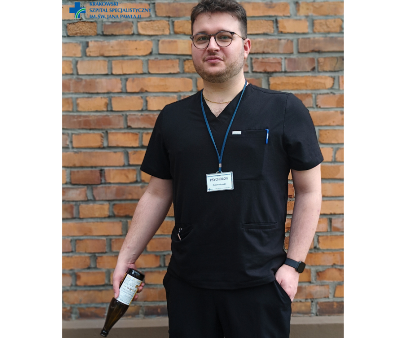 Piotr Rutkowski psycholog na tle ceglanej ściany pozujący z butelką w ręce w czarnym stroju.