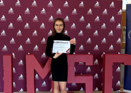 Studentka Julia Kościanek trzymająca w rękach przyznany certyfikat na tle bordowej fotościanki z logotypem IMSC.