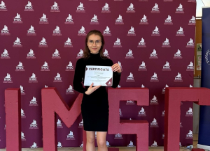 Studentka Julia Kościanek trzymająca w rękach przyznany certyfikat na tle bordowej fotościanki z logotypem IMSC.