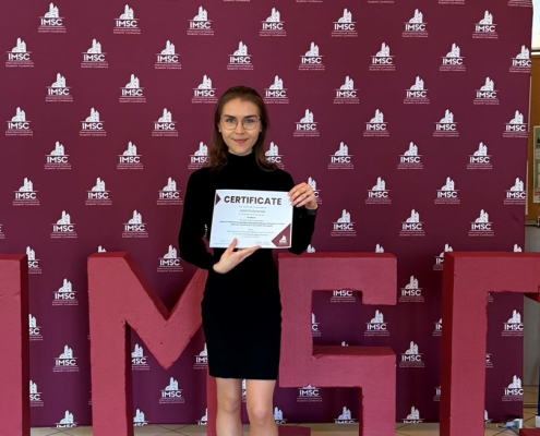 Studentka Julia Kościanek trzymająca w rękach przyznany certyfikat na tle bordowej fotościanki z logotypem IMSC.