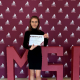 Studentka Julia Kościanek trzymająca w rękach przyznany certyfikat na tle bordowej fotościanki z logotypem IMSC.
