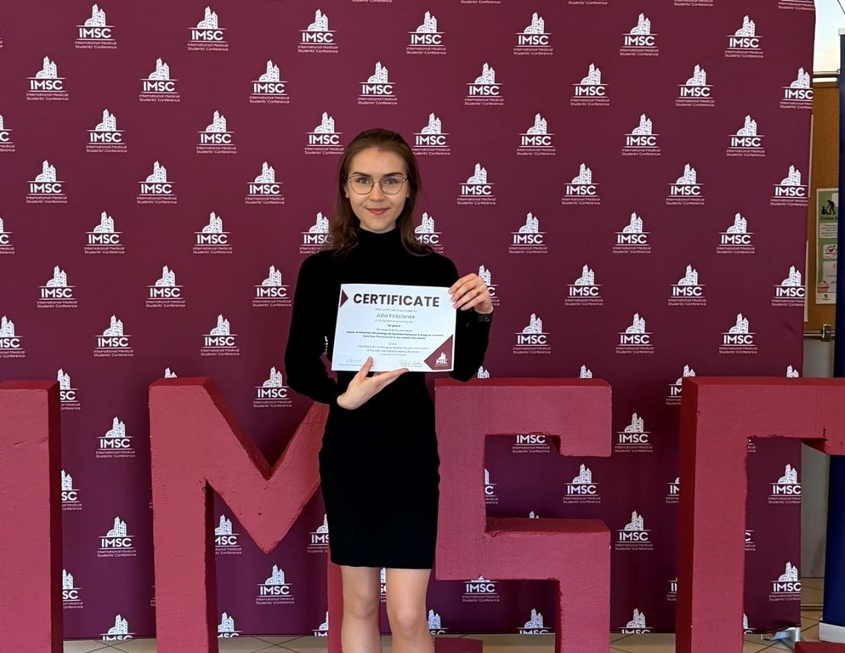 Studentka Julia Kościanek trzymająca w rękach przyznany certyfikat na tle bordowej fotościanki z logotypem IMSC.